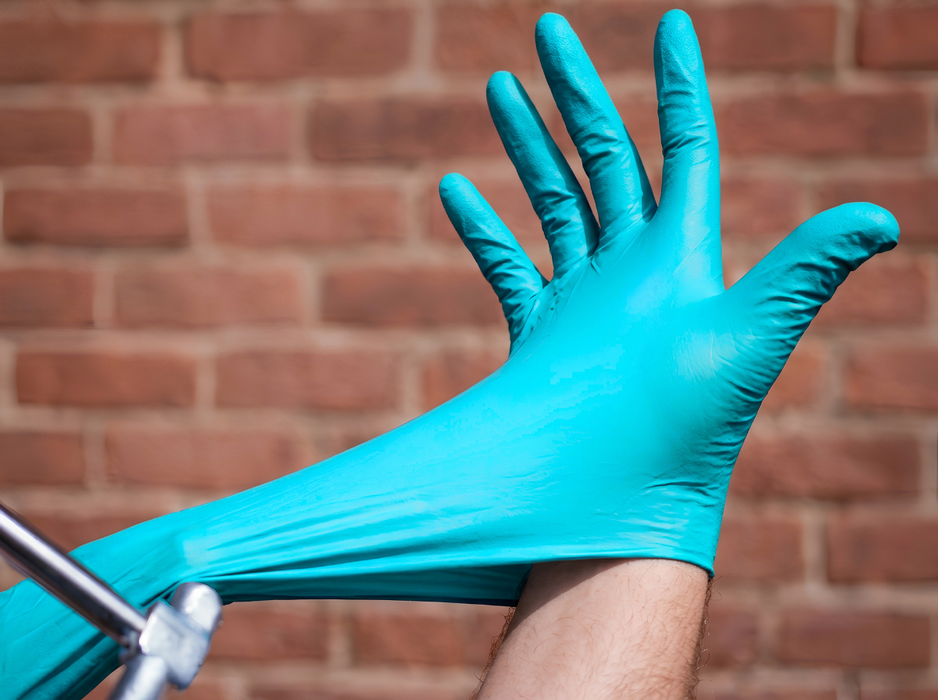 Hand putting on Abena Blue 5 Mil Nitrile Exam Glove against a brick wall background, illustrating durable fit, comfort, and protective barrier for medical, laboratory, and cleaning applications